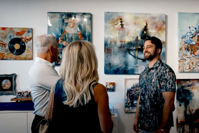 Man stands in gallery in front of paintings with collectors